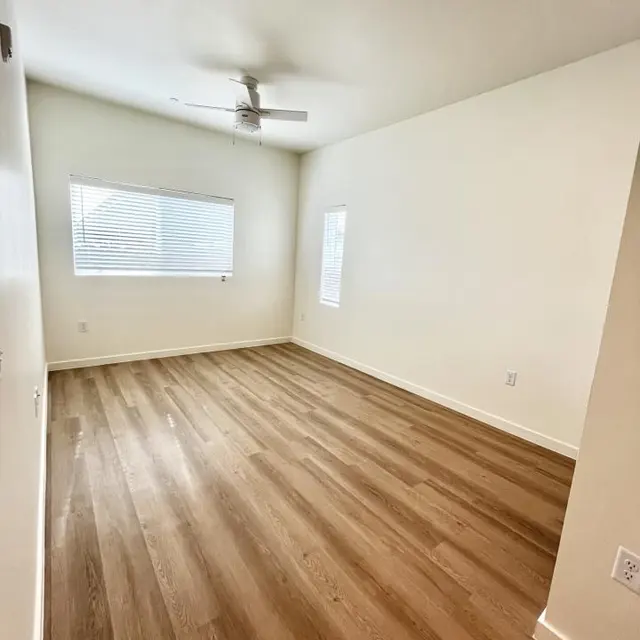 An empty room with wooden flooring and two windows, one of which has blinds.