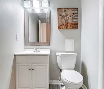 A modern bathroom featuring a white cabinet sink, a toilet, and a large mirror with three light fixtures above it. The walls are painted light gray, and there is a piece of artwork hung above the sink.