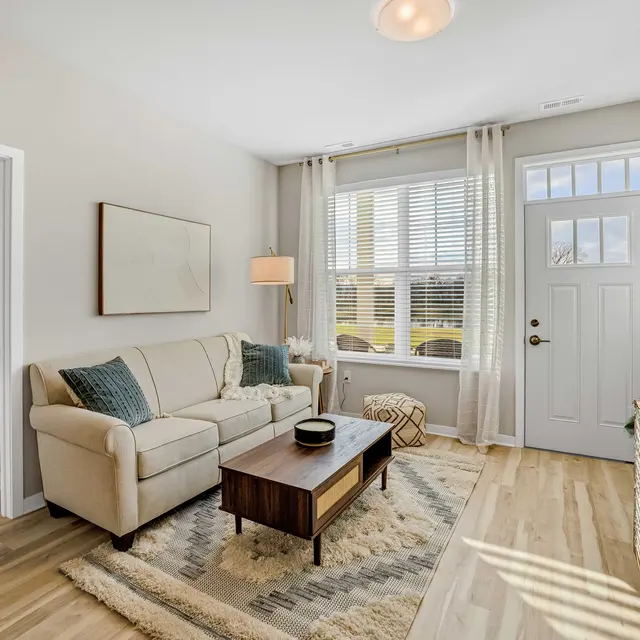 A cozy living room featuring a sofa, coffee table, and large windows with natural light. There is a doorway leading to another room, creating a welcoming atmosphere.