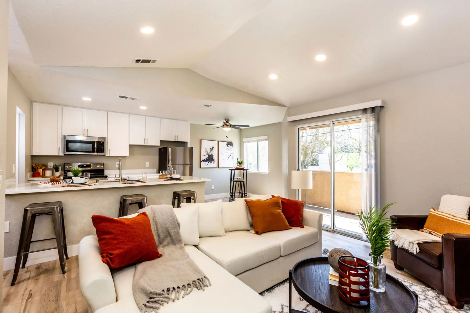 A modern living room featuring a white sectional sofa adorned with colorful pillows, a round coffee table, and large windows leading to a balcony. The adjacent kitchen includes white cabinets, stainless steel appliances, and a casual dining area.