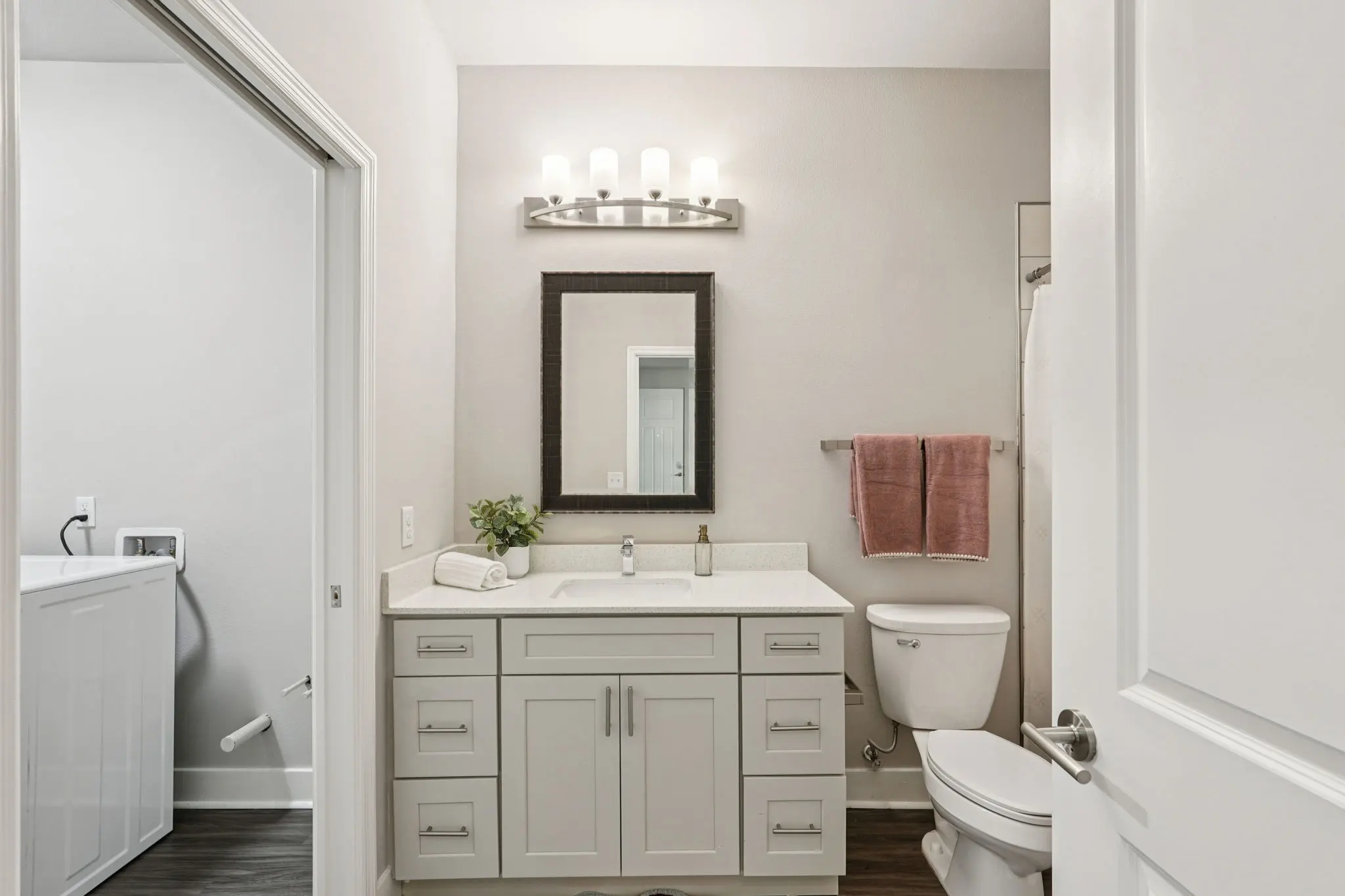 A modern bathroom featuring a double sink vanity with gray cabinetry, a large mirror, a framed mirror above the sink, a white toilet, and a pink towel hanging on the wall. The room is brightly lit, with a washer and dryer visible in the corner.