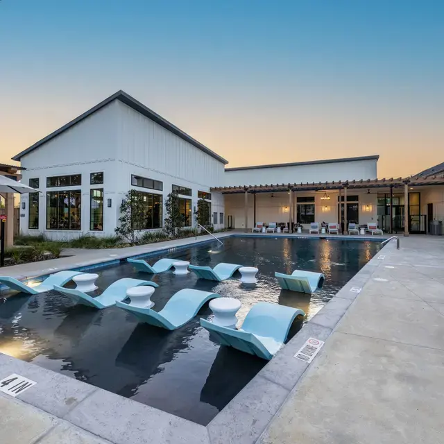 A modern pool area featuring a sleek design with wave-like lounge chairs partially submerged in water. Surrounding the pool are well-manicured plants and an elegant building with large windows in the background. The sky is clear with hints of sunset colors.