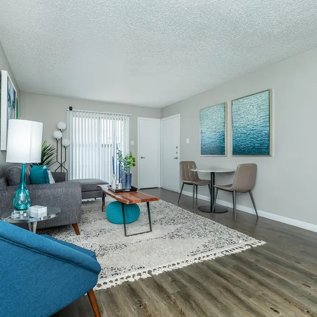 A modern living room featuring a gray sofa, a blue accent chair, a coffee table, and a dining table with two chairs. Large windows with blinds bring in natural light. Decorative wall art in blue and green tones adorns the walls, and a rug lies on the floor.
