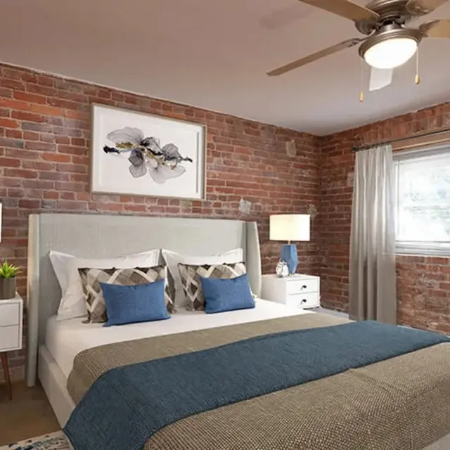 A modern bedroom with exposed brick walls, featuring a queen bed with decorative pillows, two bedside tables with lamps, and a piece of artwork on the wall.