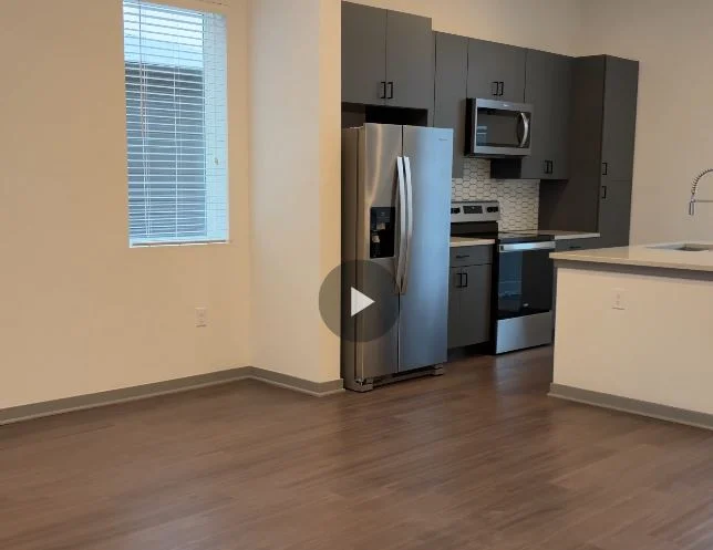 A modern kitchen and living area in an apartment, featuring stainless steel appliances, gray cabinetry, and large windows allowing natural light.