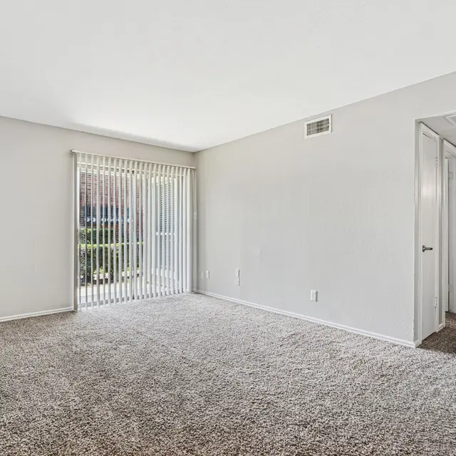 A spacious, empty living room with light grey walls and carpet. There is a large sliding glass door with vertical blinds on one side, leading to an outdoor area. A doorway on the right leads to another room, while the overall atmosphere is bright and uncluttered.