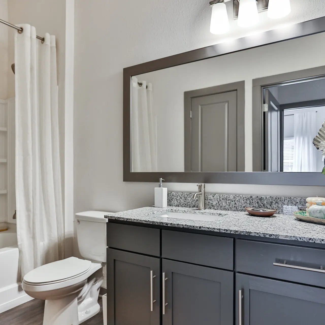A modern bathroom featuring a sleek vanity with granite countertop, a large mirror, a white bathtub with a shower curtain, and tasteful decor, including a small plant and neatly arranged toiletries.