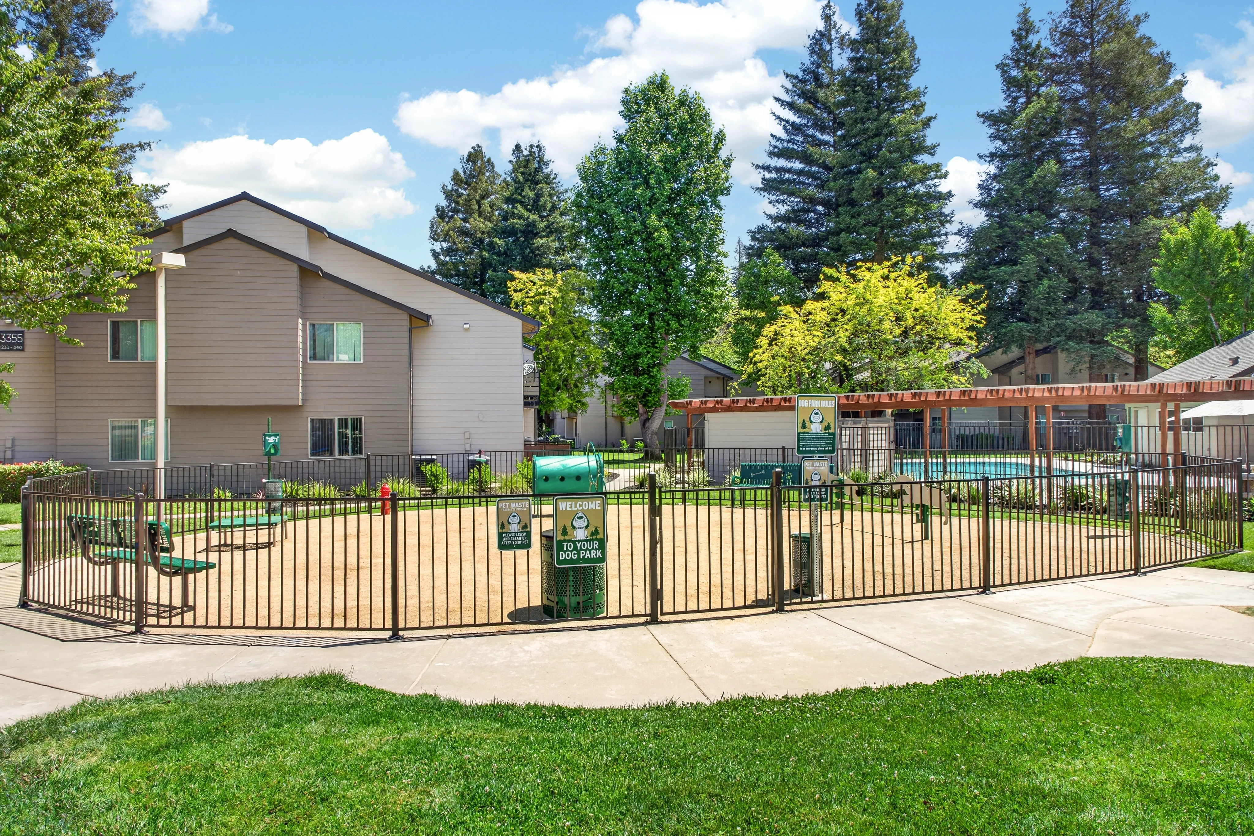 A fenced playground area surrounded by green grass, with two apartment buildings visible in the background and several trees nearby, under a blue sky with scattered clouds.