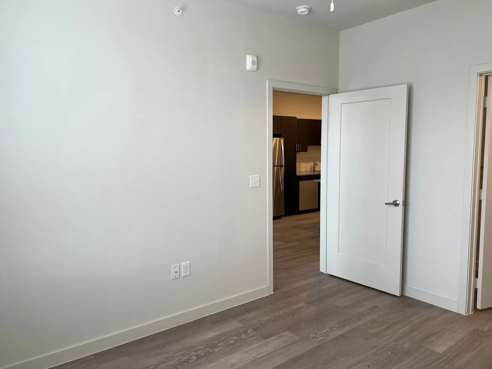 Empty Apartment Room An empty room with light-colored walls and wood flooring, featuring an open door leading to a kitchen area in the background.