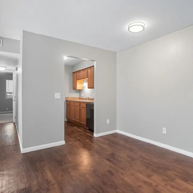 A spacious modern living area with wooden flooring, light gray walls, and a kitchen partly visible. The area is well-lit by a ceiling light, creating a warm ambiance.