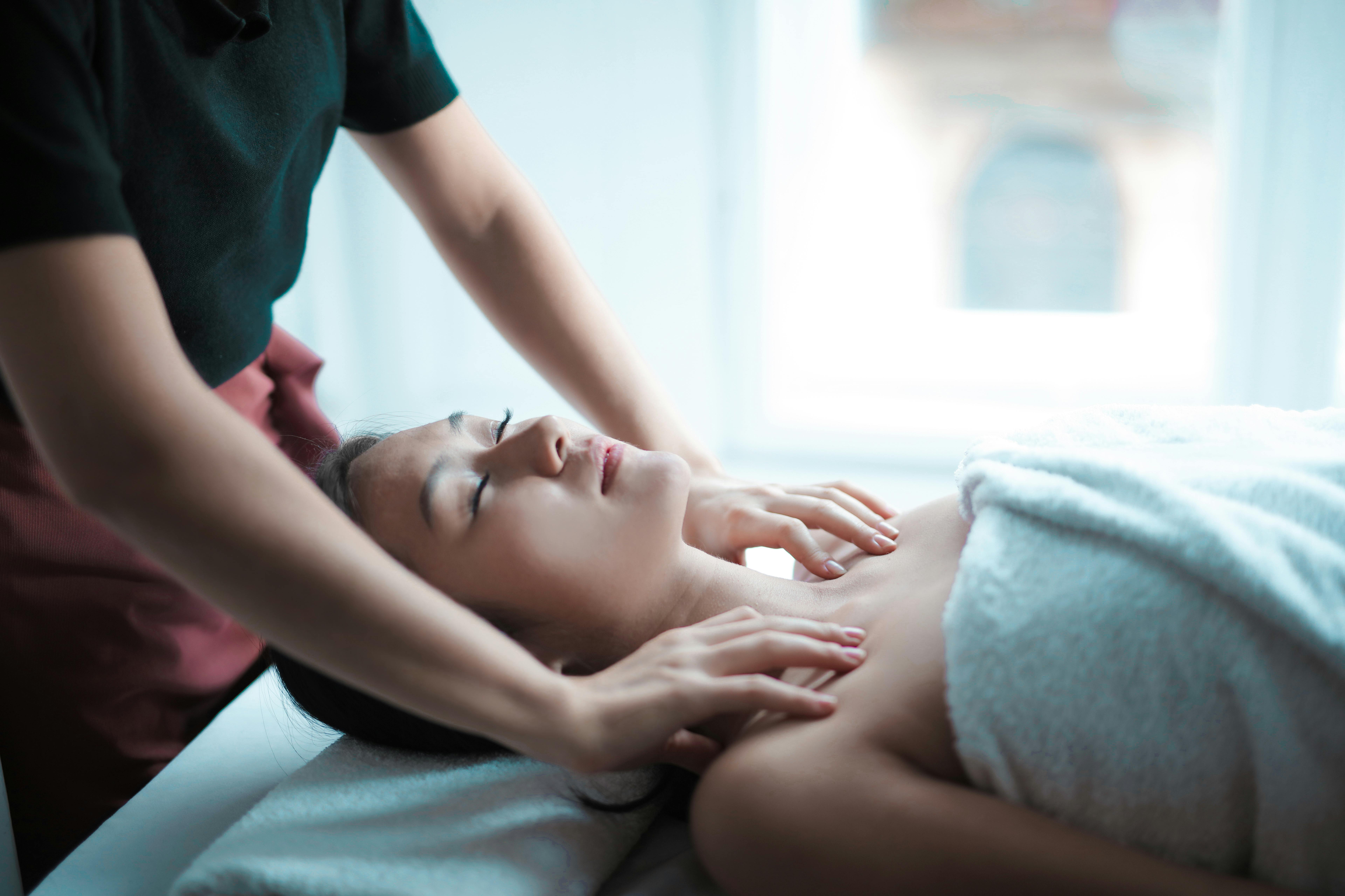 Spa Massage Session A person receiving a massage, lying on a treatment table. The masseuse, wearing a dark shirt, is gently massaging the person's neck and shoulders. The recipient appears relaxed and is covered with a white towel.