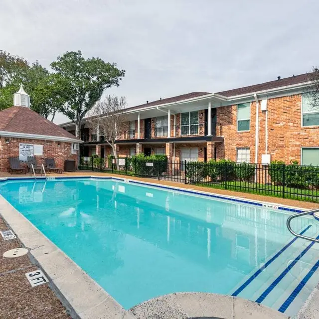 A clean, clear swimming pool in an apartment complex, surrounded by a stone patio and chairs, with a gazebo and landscaped greenery in the background.