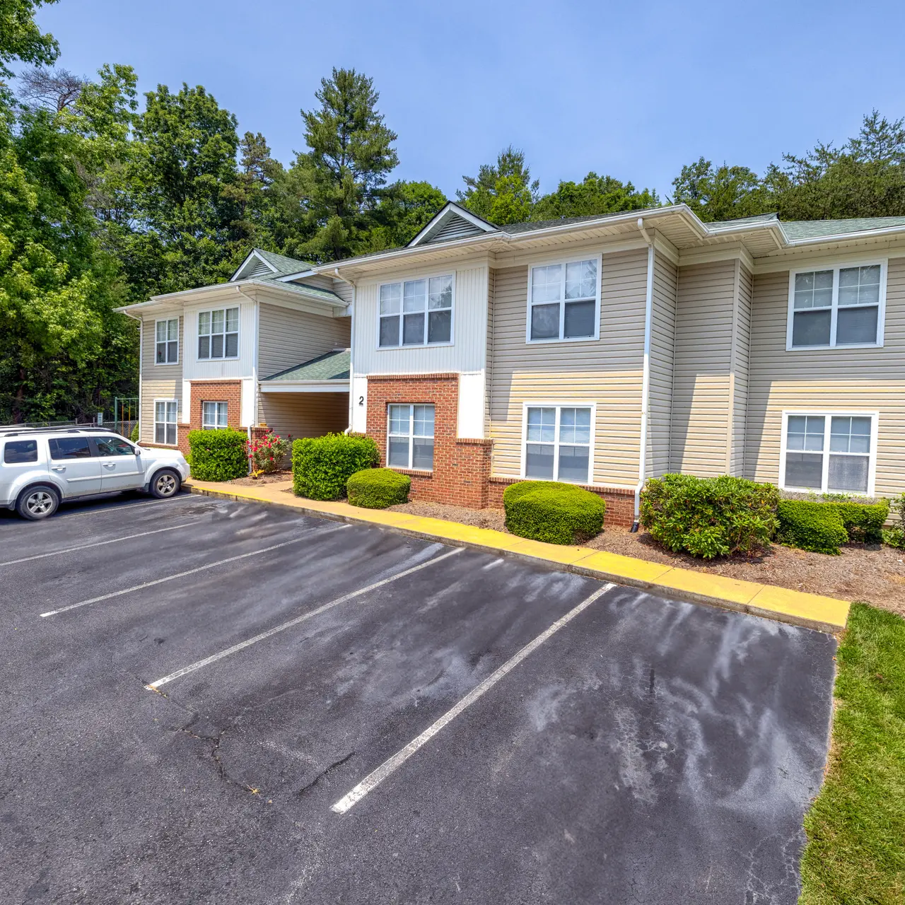 A two-story apartment building surrounded by greenery, with a large parking area in front.