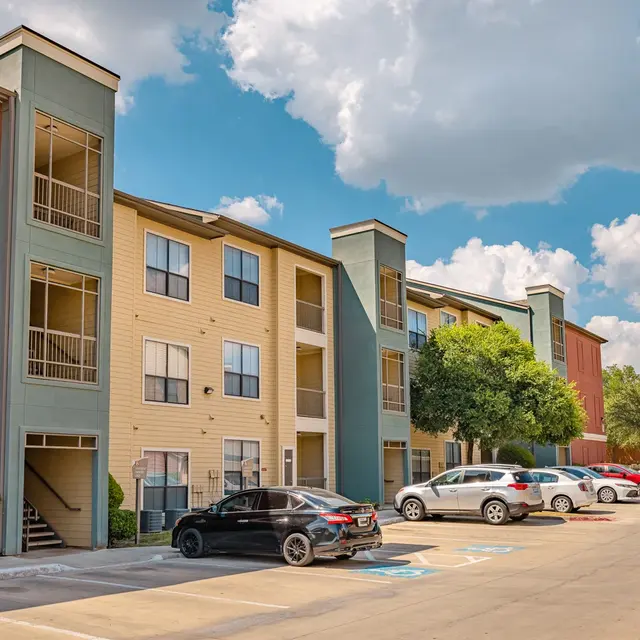 A view of an apartment complex with multiple stories, featuring balconies and a parking lot with vehicles. The sky is partly cloudy and a tree is visible in front of the building.