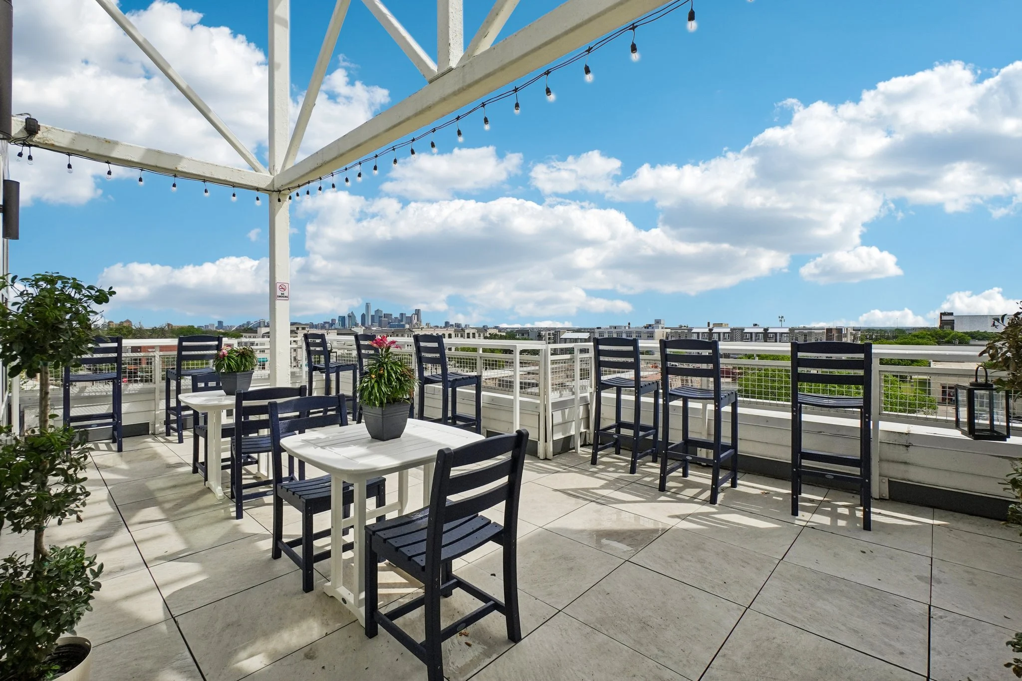Rooftop Cafe with City Skyline A rooftop cafe with outdoor seating, featuring tall chairs and small tables. The area has potted plants and a clear view of the skyline under a blue sky with scattered clouds.