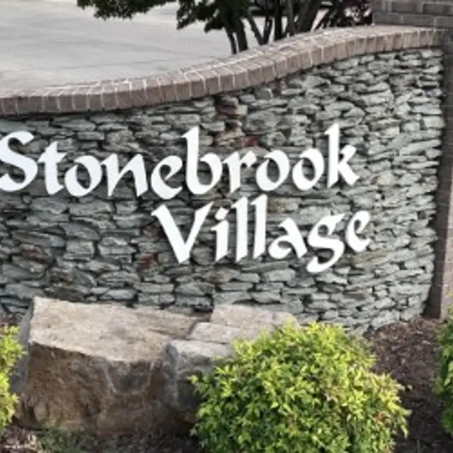 A stone and brick sign displaying the name 'Stonebrook Village' in white letters, surrounded by green bushes.