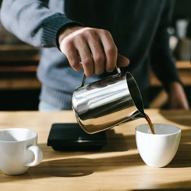 A person pouring coffee from a metal pitcher into a white cup on a wooden table, with another empty cup nearby and a plant in the foreground.