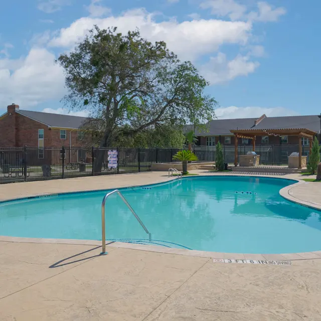 A clear, inviting swimming pool surrounded by a concrete deck, palm trees, and grassy areas, with a few houses in the background under a blue sky with scattered clouds.