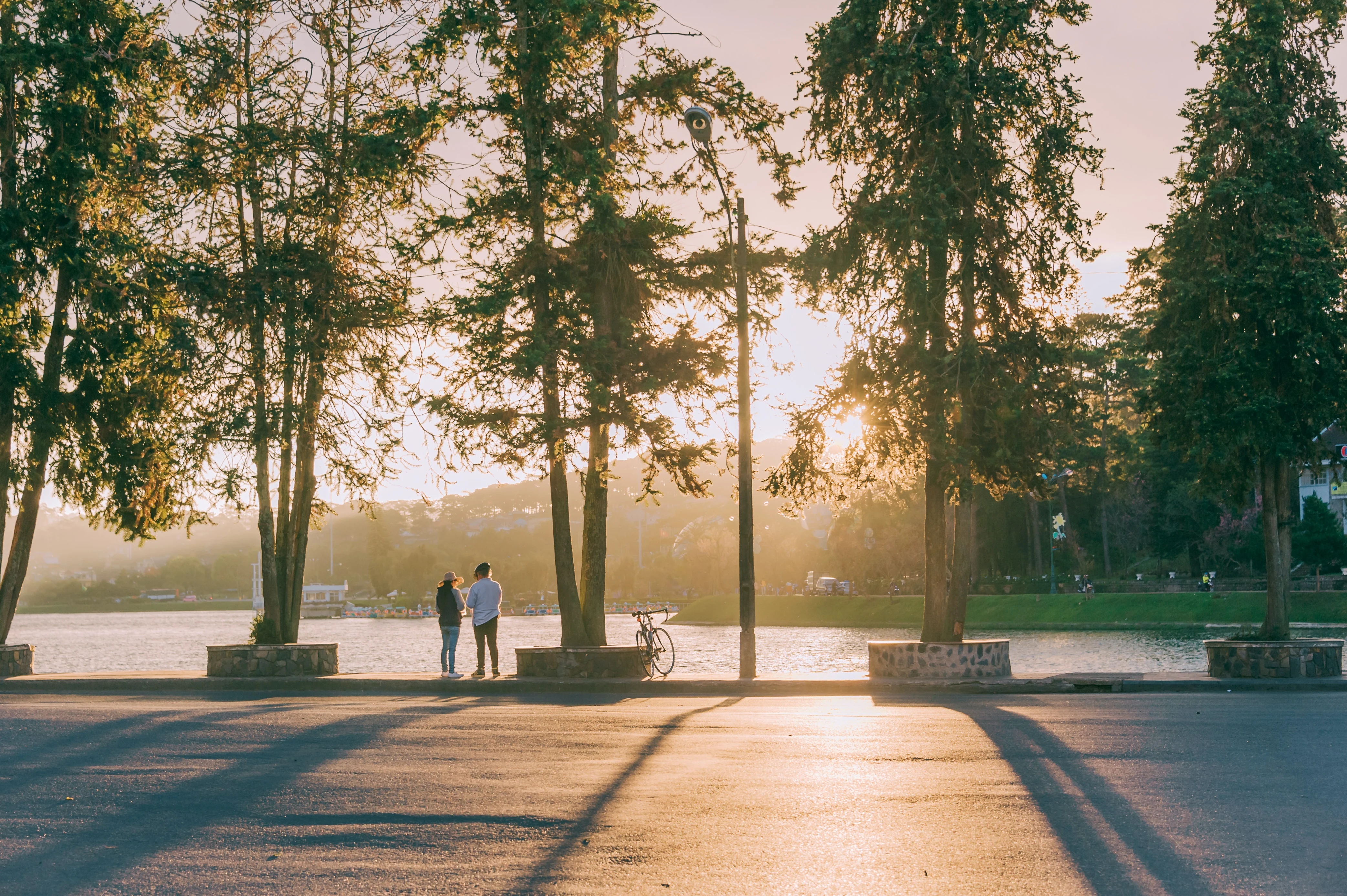 A couple standing by a lake during sunset, surrounded by tall trees casting long shadows. A bicycle rests nearby on the pavement.