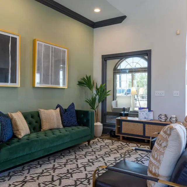 A stylish modern living room featuring a green velvet sofa with decorative pillows, two black armchairs, and a patterned rug. The walls are painted in a soft green hue, adorned with two abstract framed artworks. A plant is positioned near a large window that provides natural light.