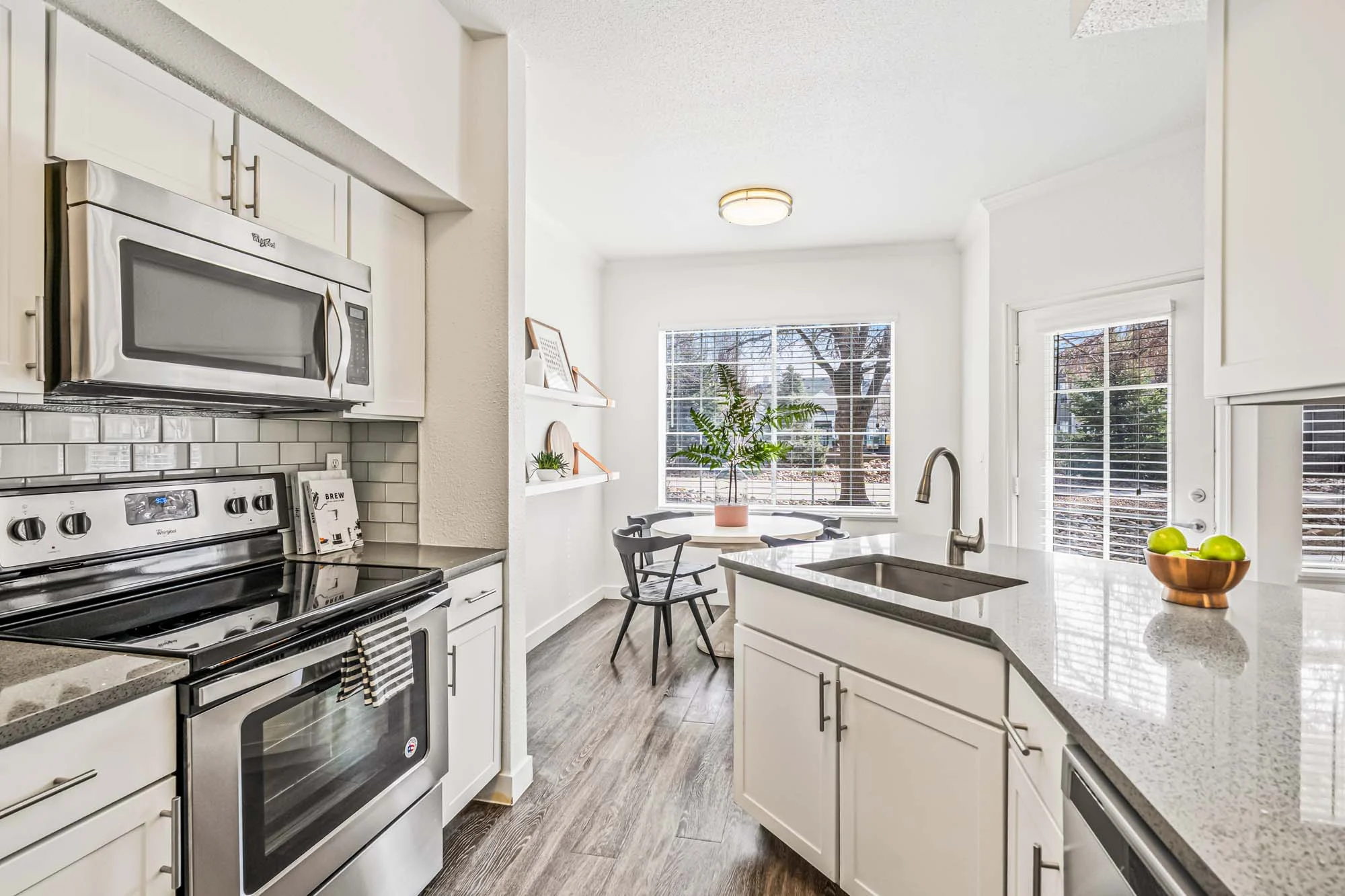 A modern kitchen featuring stainless steel appliances, a gray tile backsplash, and white cabinetry. The kitchen includes a sink with a view of a small dining area bathed in natural light from a window.