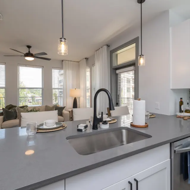 Modern Kitchen and Living Room A modern kitchen with a countertop overlooking a cozy living room. The kitchen features white cabinets, a gray countertop, and pendant lighting. The living room has a light-colored sofa, patterned rug, and large windows with sheer curtains.