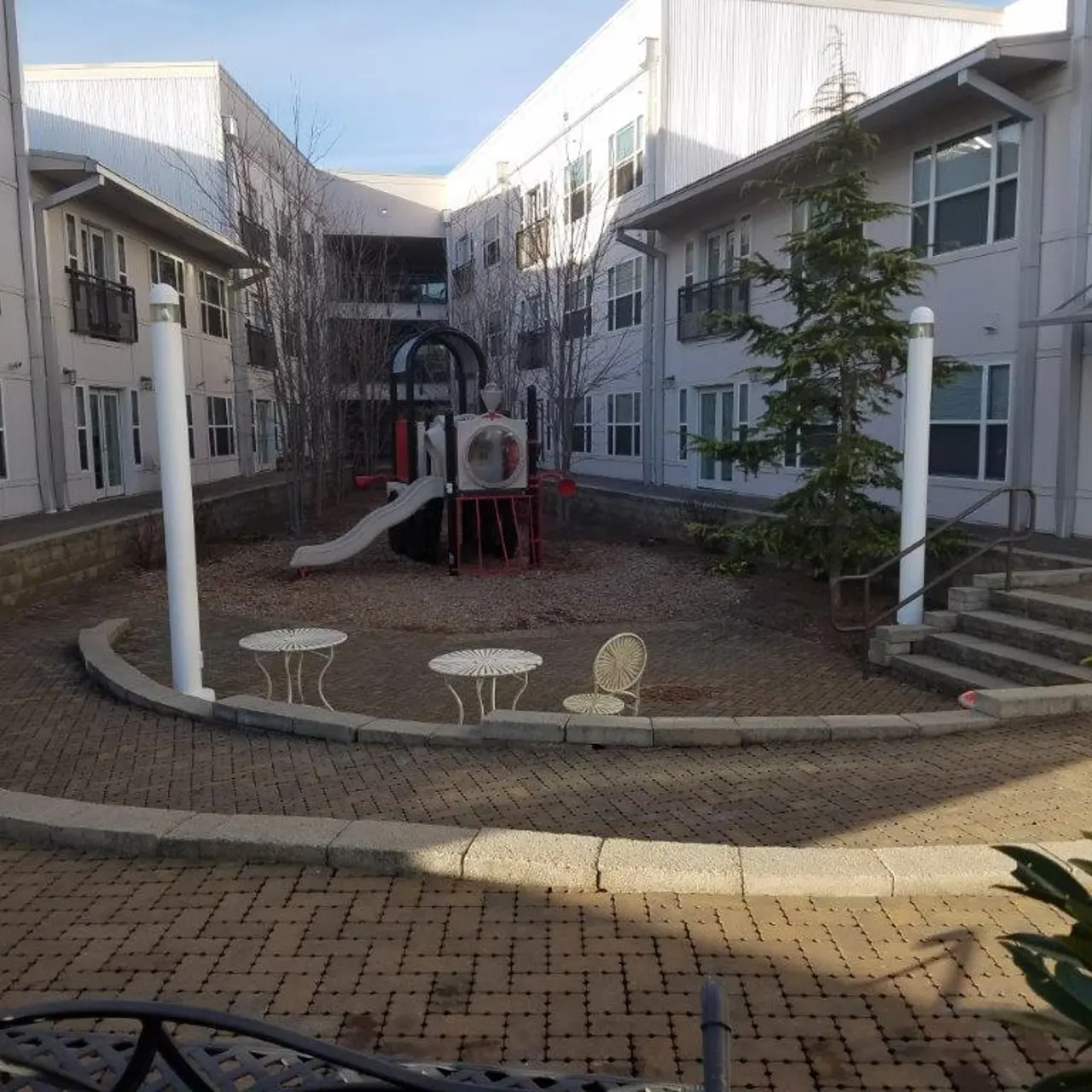 A playground area in a courtyard surrounded by apartment buildings. The playground features a small slide and play structure, with a few white chairs placed on a patterned stone pathway. There are some trees and a quiet, empty atmosphere.