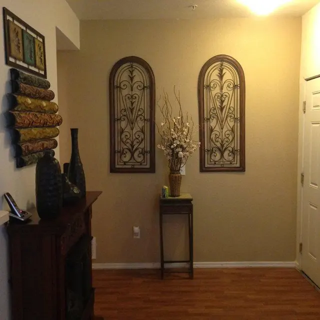 Decorative Entryway Interior A cozy entryway featuring decorative items. Two ornate wall hangings are positioned above a small table with a flower arrangement. A dark vase decorates the left side, and a textured wall piece is to the left on a shelf. The floor is wood, and there's a door leading outside.