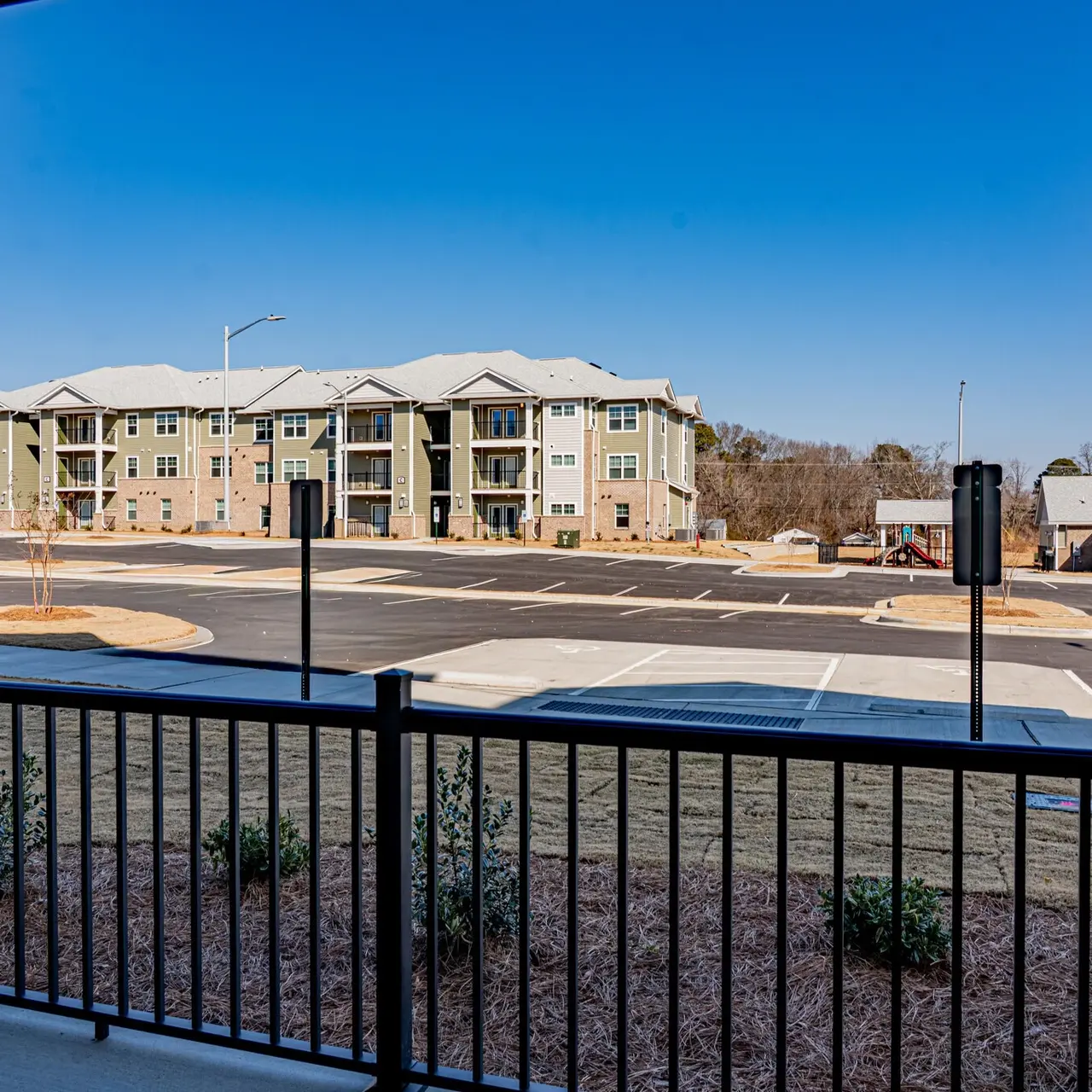 View from a balcony overlooking a parking lot and adjacent apartment building under a clear blue sky.