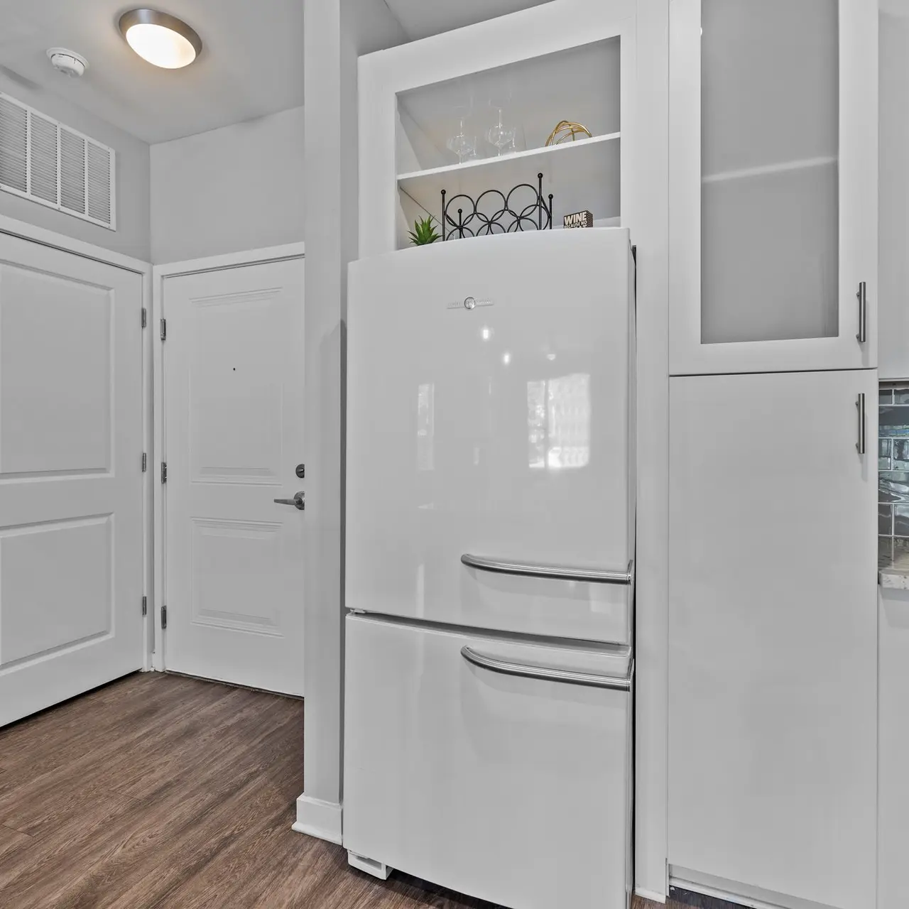 A bright and modern kitchen featuring a white refrigerator, light wood flooring, and an open entrance. There are glass cabinet doors and a tiled backsplash by the stove.