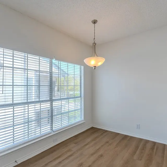 A bright dining area featuring a window with vertical blinds, light wood flooring, and a pendant light hanging from the ceiling.