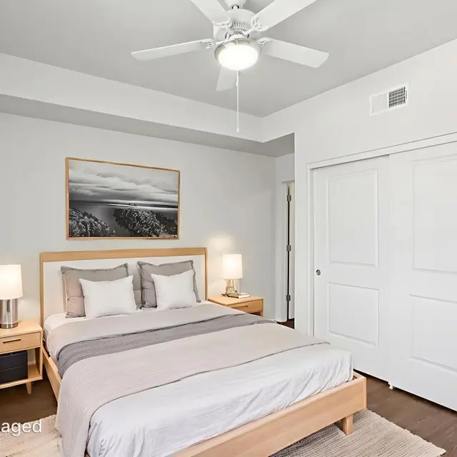 Modern Bedroom Interior A modern bedroom featuring a large bed with gray and white bedding, two bedside tables with lamps, a piece of artwork on the wall, and a ceiling fan. The room has light-colored walls and wooden flooring.