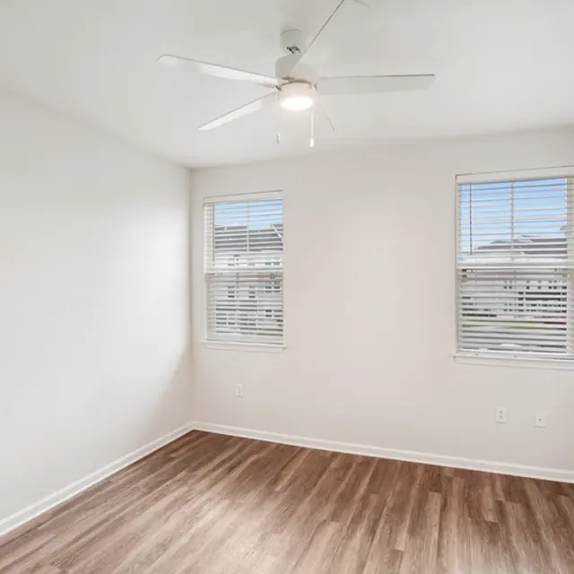 A well-lit empty room with light walls, a ceiling fan, two windows, and wooden flooring.
