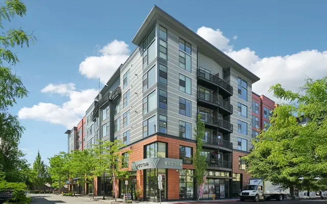 A modern apartment building featuring multiple floors with a mix of colored facades and large windows, surrounded by lush green trees and a clear blue sky.