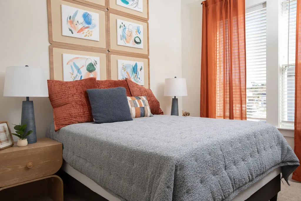 A cozy bedroom showcasing a neatly made bed with gray bedding and colorful decorative pillows, two white lamps on either side, and a pair of large orange curtains filtering sunlight through the window.