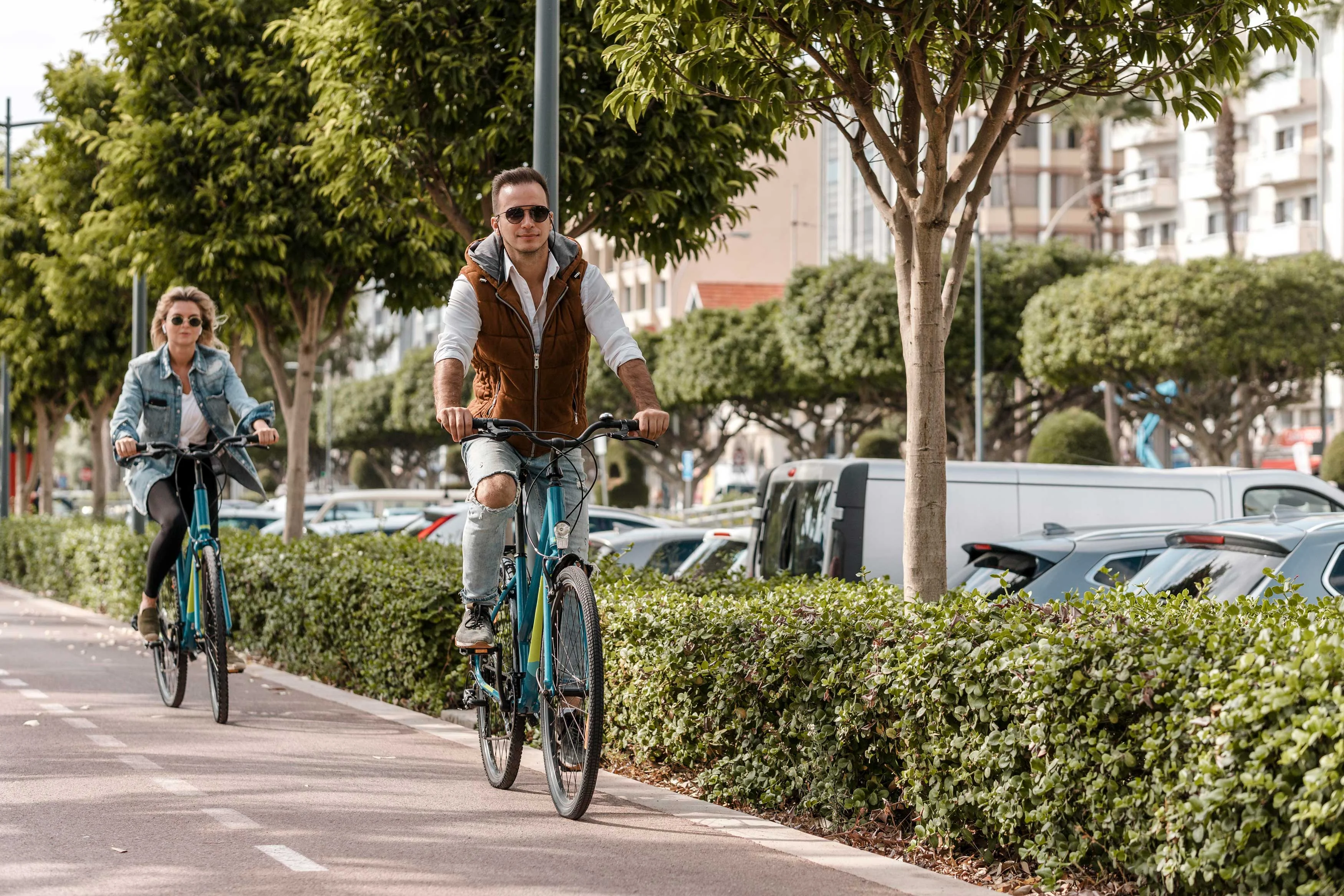 A man and woman riding bicycles on a path surrounded by lush greenery and parked cars.