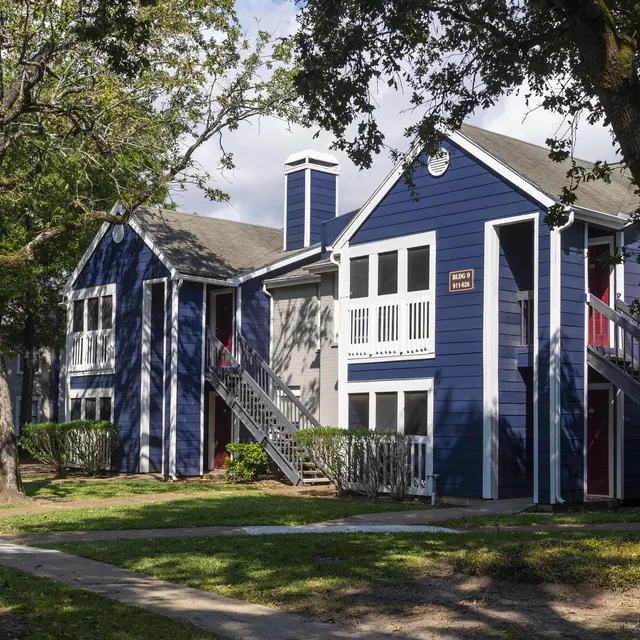 A blue and gray apartment complex with green lawns and trees surrounding it, featuring balconies and stairs for access.