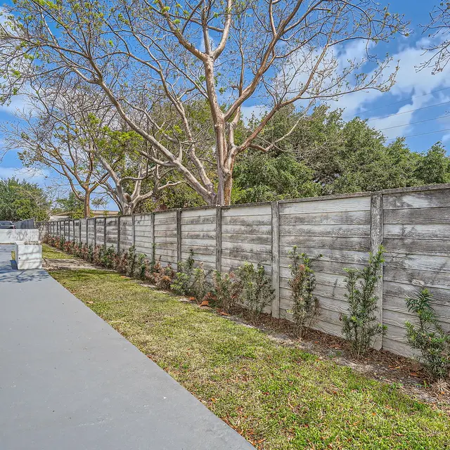 A view of a narrow outdoor pathway beside a wooden fence, adorned with small shrubs and trees in the background under a bright blue sky.