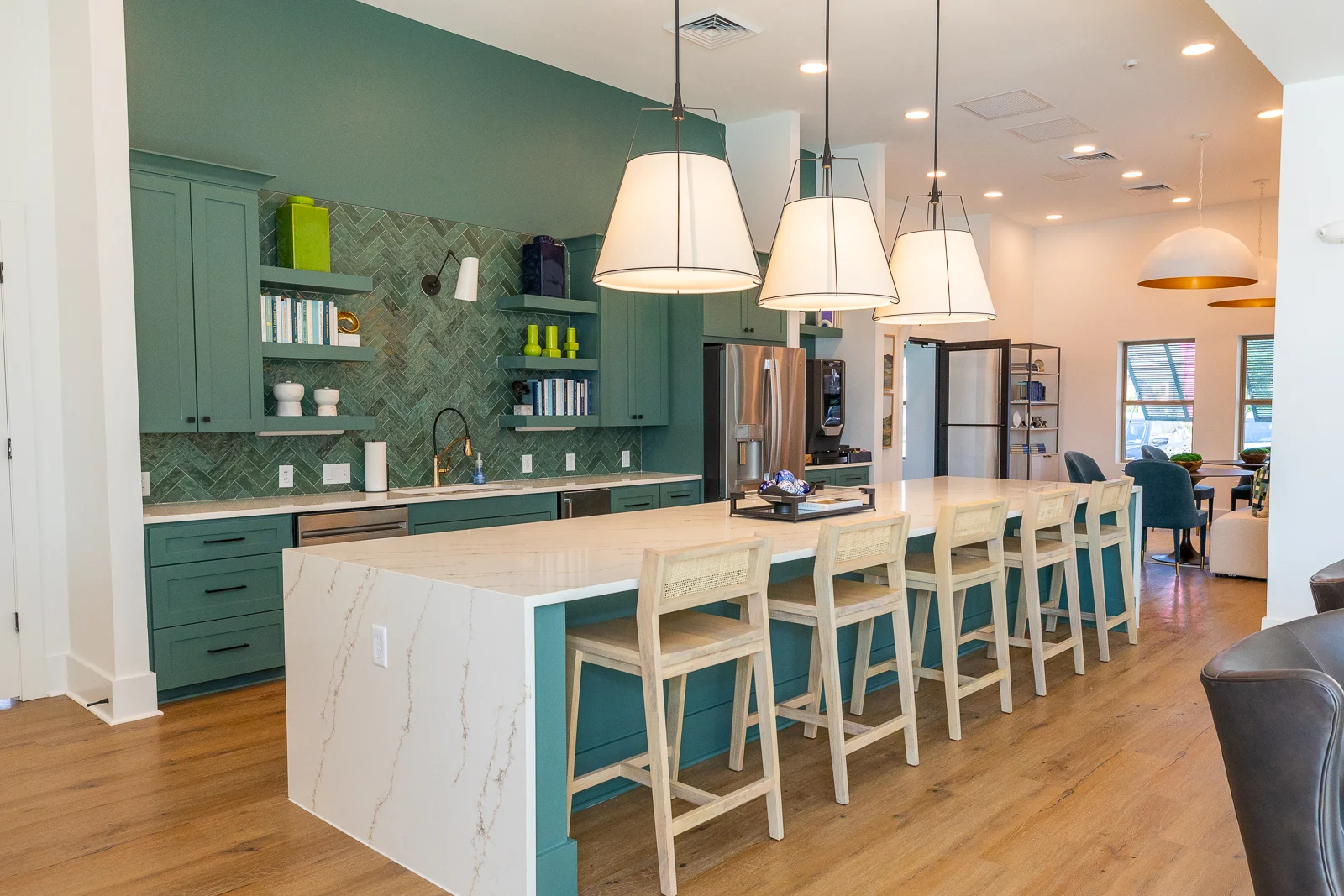 A modern kitchen featuring a long island with bar stools, teal cabinets, a herringbone backsplash, and pendant lighting. The kitchen has stainless steel appliances and wooden flooring.