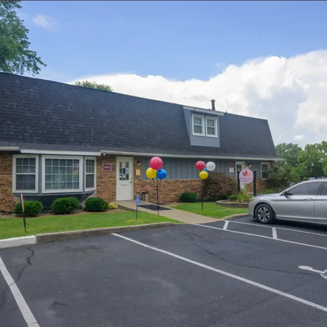 A brick building with a sloped roof, featuring colorful balloons in front, surrounded by green grass and parked cars.