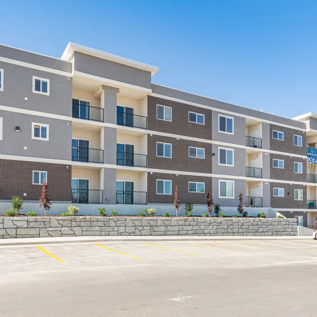 A modern four-story apartment building with a mix of gray and brown exterior, featuring balconies on each level and a parking lot in front.