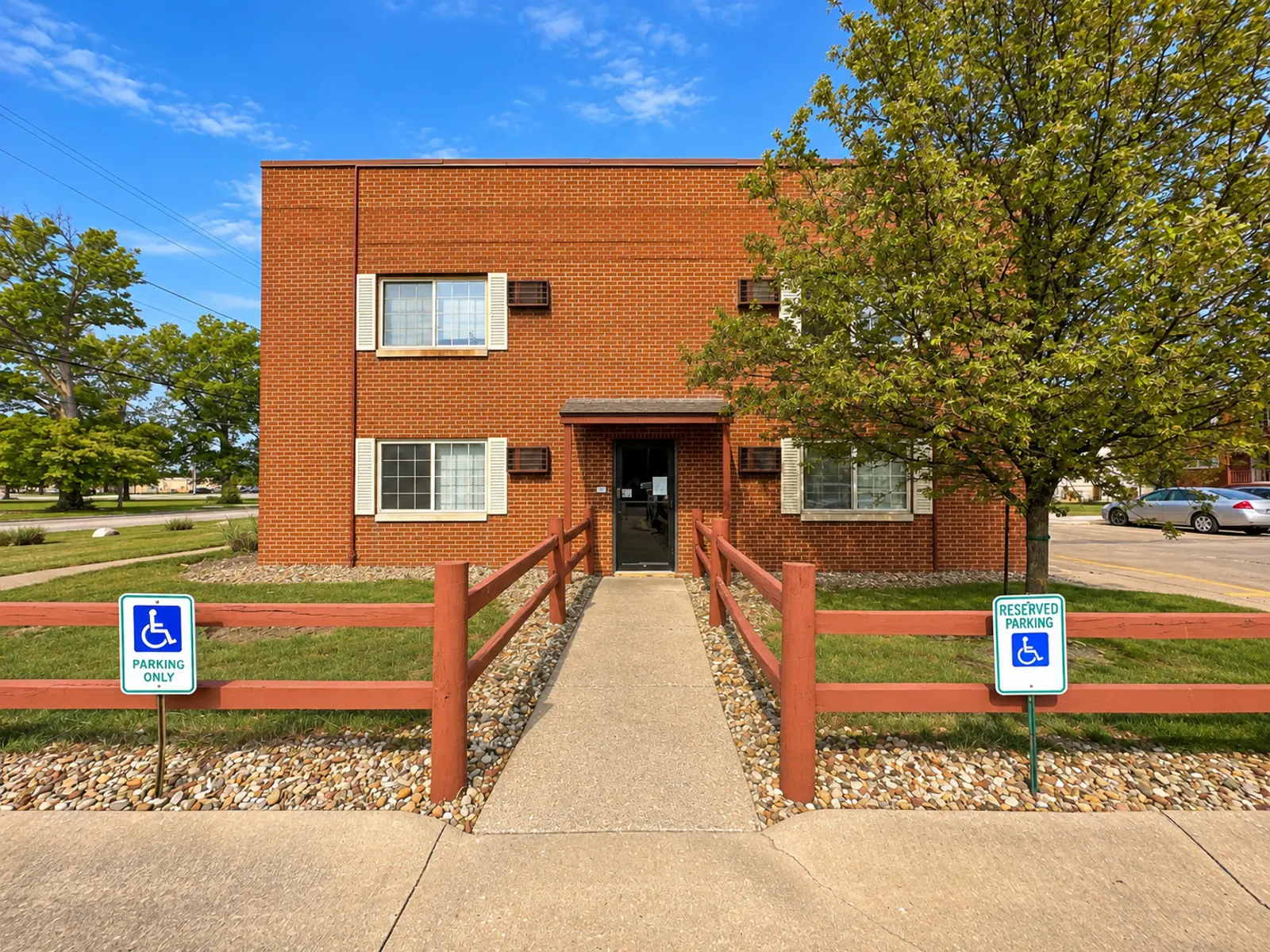 Exterior view of a two-story brick apartment building with a pathway leading to the entrance and accessible parking signs.