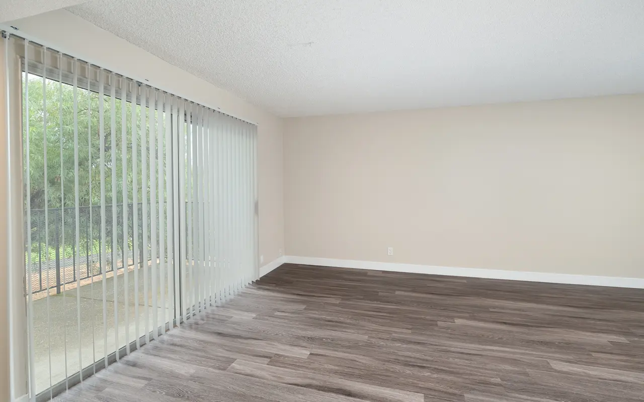 An empty room with beige walls and wooden flooring, featuring large sliding glass doors leading to a patio with greenery outside. The room is well-lit and spacious, creating a minimalist ambiance.