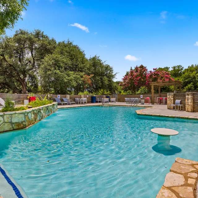 A beautiful pool area featuring clear water, stone walls, and lounge chairs surrounded by trees and greenery.