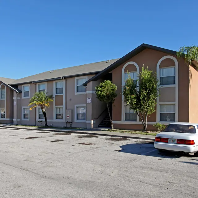 A view of a two-story apartment building surrounded by palm trees, with a parking area in front and a clear blue sky.