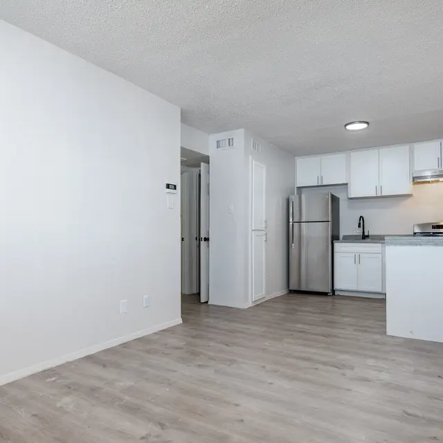 A modern apartment interior featuring a spacious living area leading to an open kitchen. The kitchen has white cabinets, stainless steel appliances, and a countertop. The flooring is light wood, and the walls are painted white, giving it a clean and bright appearance.