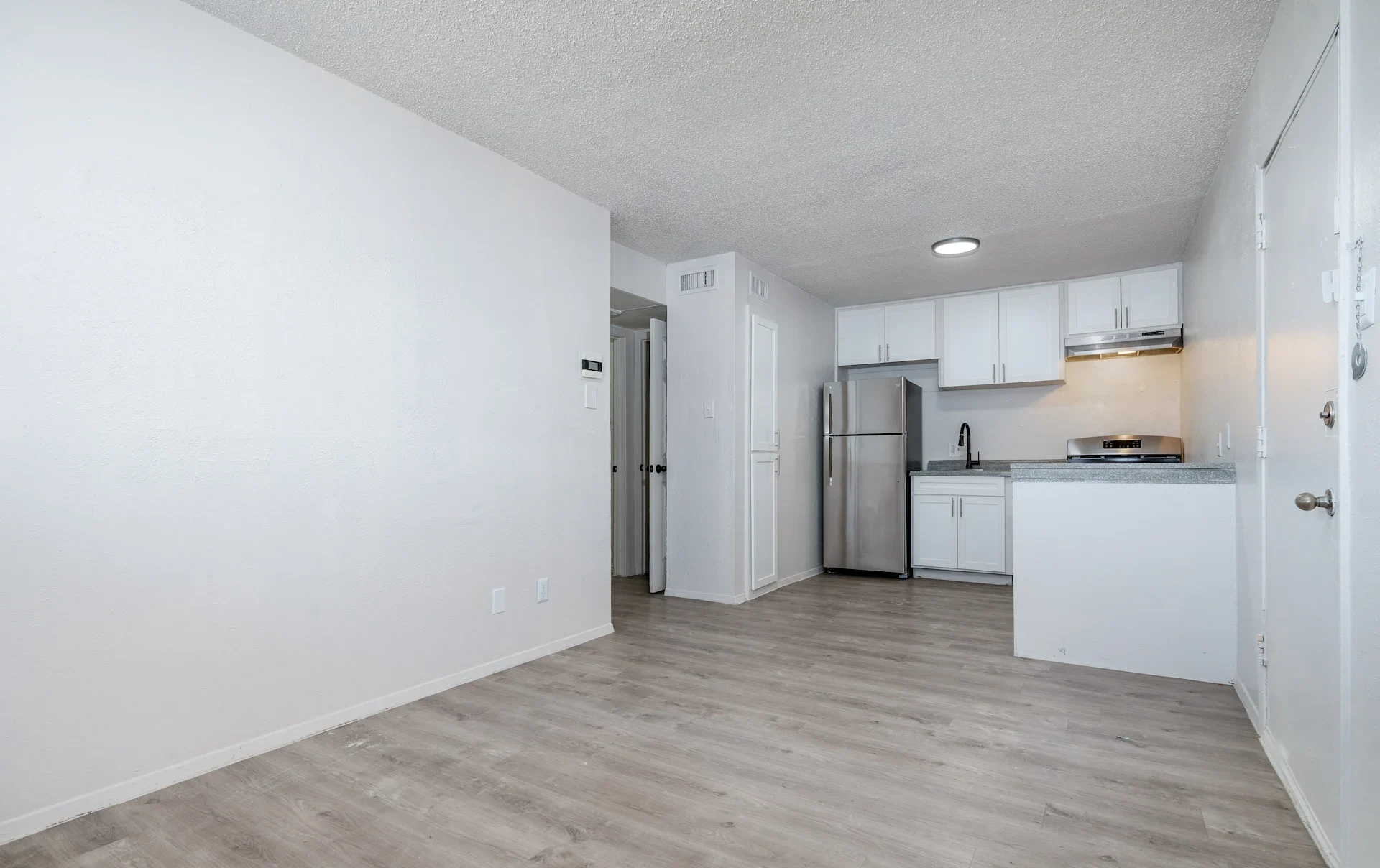 A modern apartment interior featuring a spacious living area leading to an open kitchen. The kitchen has white cabinets, stainless steel appliances, and a countertop. The flooring is light wood, and the walls are painted white, giving it a clean and bright appearance.