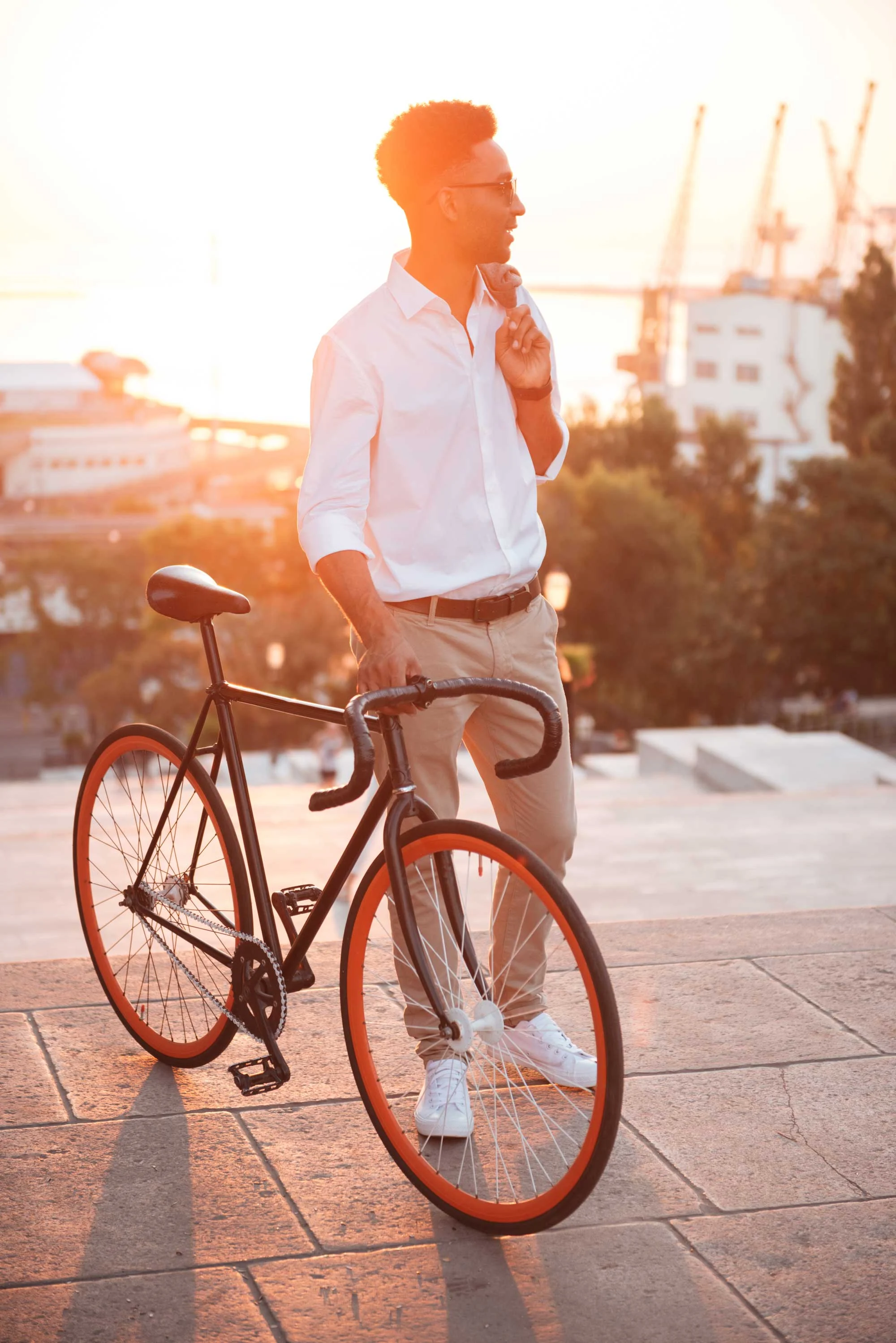 A man standing beside a black bicycle during sunset, wearing a white shirt and beige pants, with a cityscape in the background.