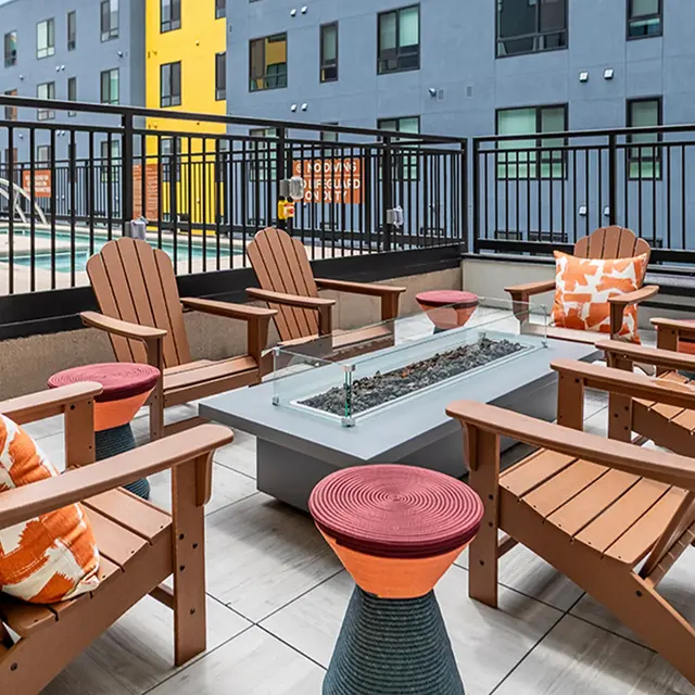 A rooftop lounge with wooden chairs, a fire pit, and colorful accents. The background features buildings with blue and yellow walls.