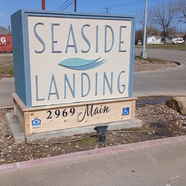 A roadside sign for Seaside Landing, displaying the name in large text with a wavy design. The sign is located at the intersection of Main Street, with a clear blue sky in the background.